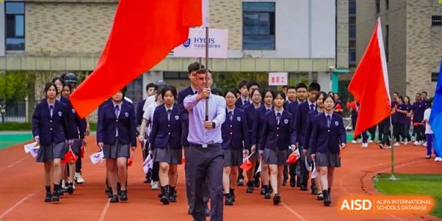 A student carrying a large red flag leads a procession of students in navy blue uniforms at Hongyi Cambridge International School (HYCIS).