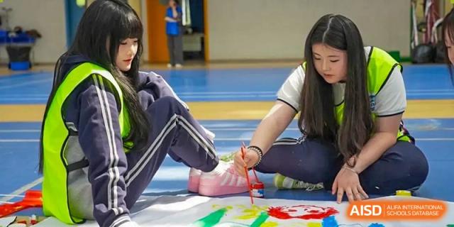 A close-up of students from Hongyi Cambridge International School (HYCIS) sitting on the gym floor, holding hands, and engaging in an indoor activity.