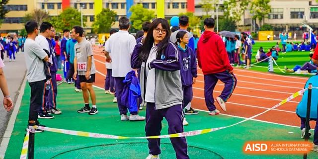 Students from Hongyi Cambridge International School (HYCIS) standing on the track wearing tracksuits and watching an event, with one student looking directly at the camera.