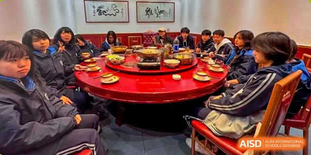 Students or staff from Hongyi Cambridge International School (HYCIS) gathered around a large, red round table for a group meal, likely hot pot or a banquet.