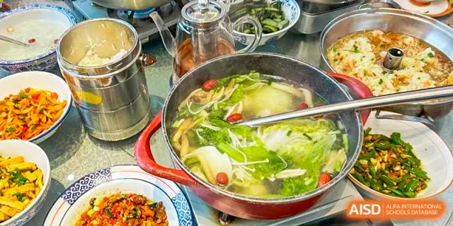A close-up of a pot of simmering hot pot or vegetable soup with leafy greens, mushrooms, and other ingredients, part of a dining scene at Hongyi Cambridge International School (HYCIS).