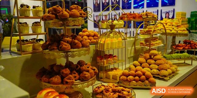 A visually appealing dessert display featuring a variety of pastries, donuts, and other baked goods stacked on tiered trays and in a gold birdcage-style stand, possibly from an event at Hongyi Cambridge International School (HYCIS).