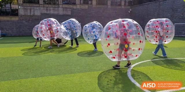Students play with large, clear inflatable bubbles on a green field at the International School of Qingdao.