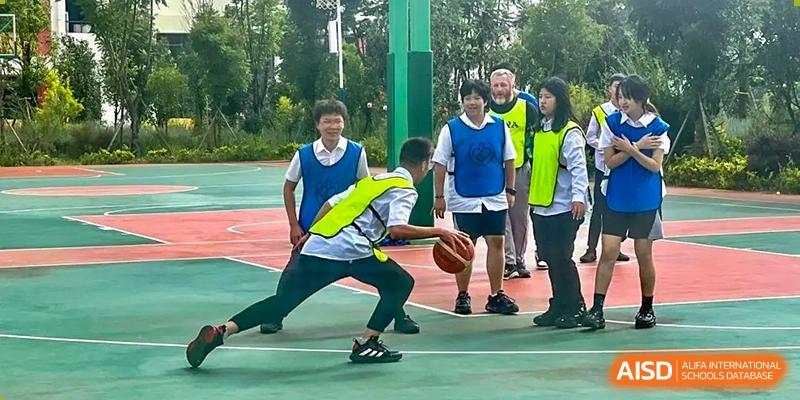 Students from Kunming World Youth Academy play a basketball game. One student in a white and blue jersey dribbles the ball while another student in a yellow safety vest defends him.