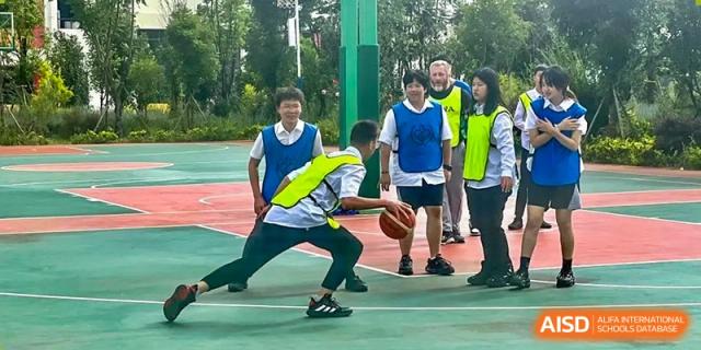 Students from Kunming World Youth Academy play a basketball game. One student in a white and blue jersey dribbles the ball while another student in a yellow safety vest defends him.