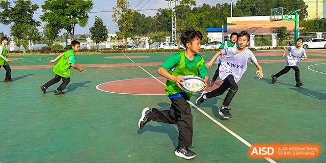 A young boy from Kunming World Youth Academy, wearing a green jersey and black pants, runs with a rugby ball on an outdoor basketball court.