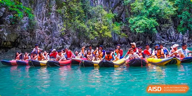A group of students from Kunming World Youth Academy smiles and poses for a photo while sitting in kayaks in a bright turquoise lagoon, surrounded by lush green cliffs.