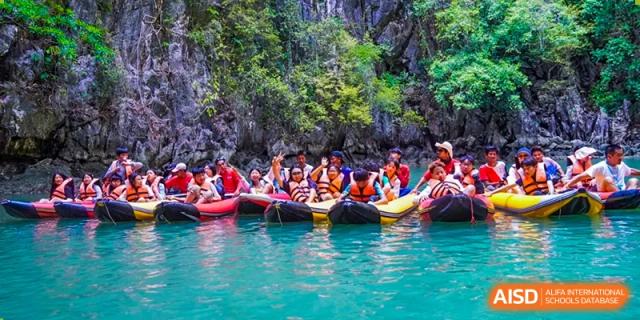 A group of students from Kunming World Youth Academy smiles and poses for a photo while sitting in kayaks in a bright turquoise lagoon, surrounded by lush green cliffs.