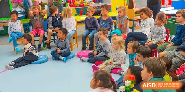 A group of young Lycée Français de Shanghai students is sitting on the floor in a circle, listening to their teacher.