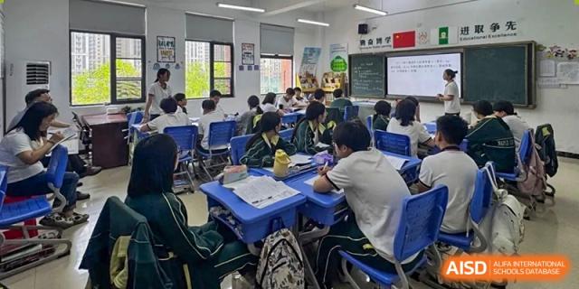 A wide shot of a classroom setting with students in green and yellow uniforms working individually at their desks, with a whiteboard visible at the front at Maple Leaf School- Xiangyang.