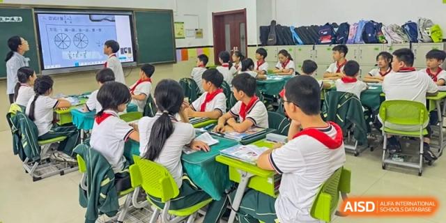 A group of students in red scarves and white shirts, sitting at green desks and focusing on classwork or a lecture at Maple Leaf School- Xiangyang.
