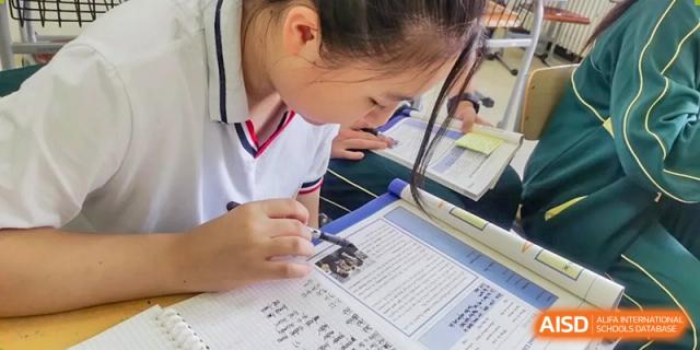 A close-up of a student in a white uniform concentrating while writing on a worksheet on their desk at Maple Leaf School- Xiangyang.