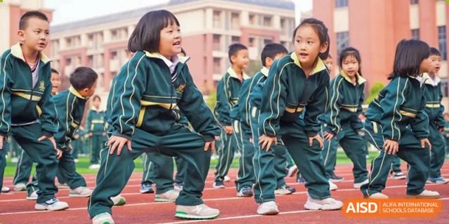 A group of young students in dark green and yellow uniforms performing an outdoor exercise or routine on a paved area at Maple Leaf School- Xiangyang.