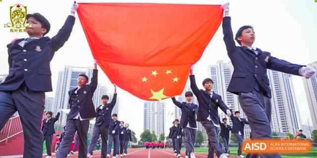 Students and faculty holding up a large Chinese flag outdoors, with a city skyline and school buildings in the background at Maple Leaf School- Xiangyang.