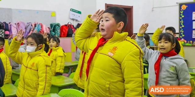 A male student in a yellow coat and red scarf standing outside and giving a salute with his hand to his forehead at Maple Leaf School- Xiangyang.