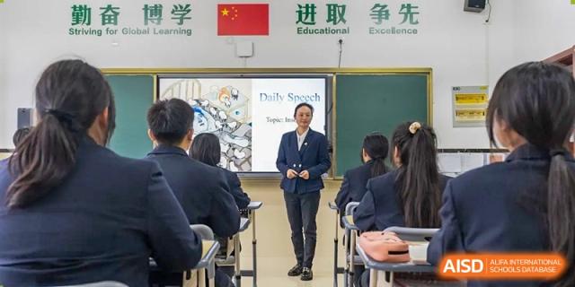 A teacher standing at the front of a classroom next to a screen, giving a presentation to seated students at Maple Leaf School- Xiangyang.