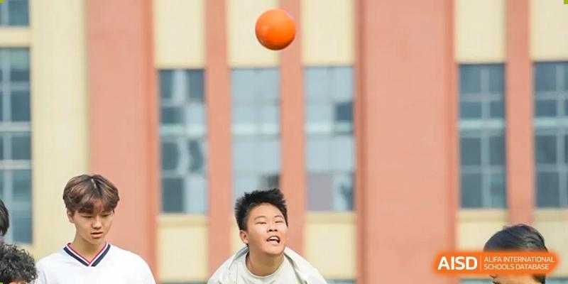 A male student from Maple Leaf School - Xiangyang looking up to catch an orange ball outdoors, with a blurred building in the background.