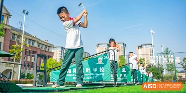A male student from Maple Leaf School - Xiangyang practicing a golf swing with a driver on a green with a school banner visible.