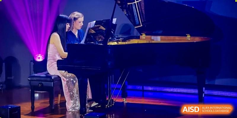 Two people, possibly students or teachers from Maple Leaf School - Xiangyang, playing a grand piano on a stage lit with blue lights.