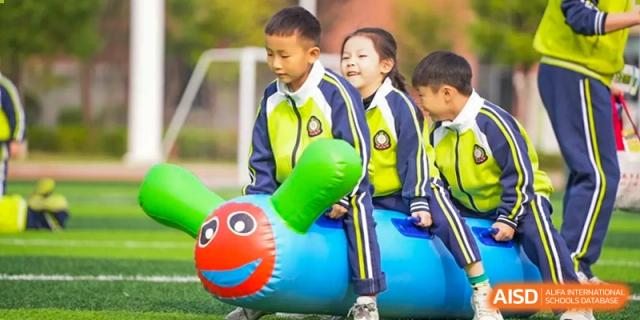 Young students from Maple Leaf School - Xiangyang smiling while sitting on a large, inflatable caterpillar play structure outdoors.