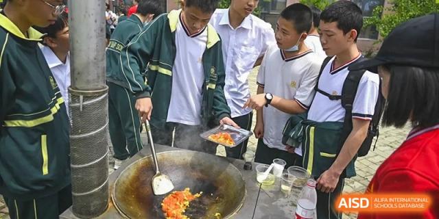 Students gathered around a large outdoor wok, watching or assisting as food is cooked, highlighting a cooking activity at Maple Leaf School- Xiangyang.