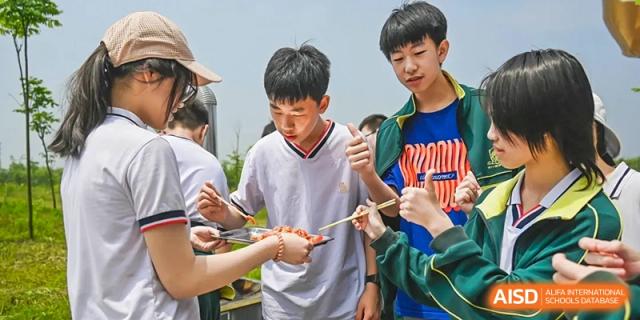 Two smiling male students receiving or taking food from a skewer during an outdoor activity at Maple Leaf School- Xiangyang.