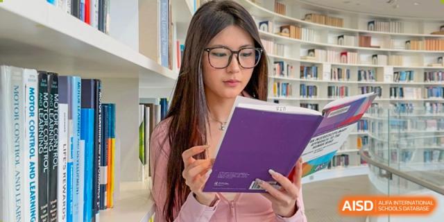 A female student wearing glasses and a pink shirt, reading a purple book in the school library at Weiming International Qingdao.