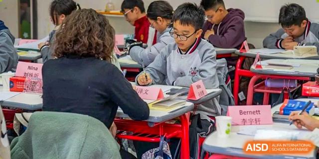 A close-up of a male student wearing a grey and red uniform vest, focused on his work during class at Weiming International Qingdao.