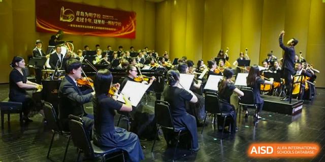 The student orchestra/band performing on stage in a yellow-lit hall. They are playing string instruments at Weiming International Qingdao.