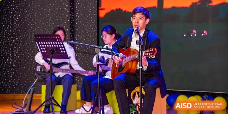A student performing on stage, playing acoustic guitar and singing into a microphone during a school event at Weiming International Qingdao.