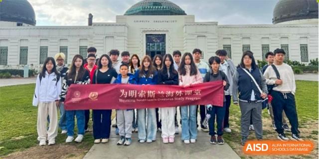 A group of students holding a red banner in front of a school building, posing for a photo at Weiming International Qingdao.