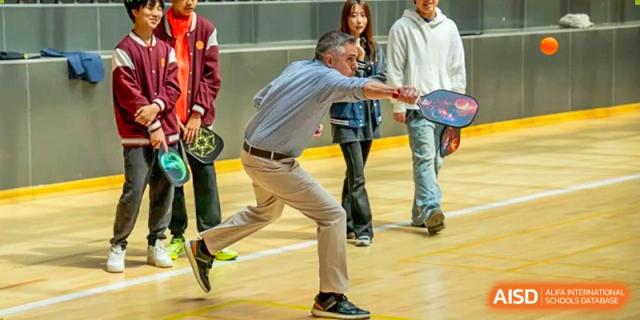 A teacher or coach leaning down to play a paddle or racket sport indoors with students watching at Weiming International Qingdao.