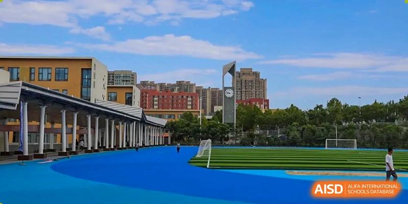 An outdoor blue running track and sports field with school buildings and a city skyline visible under a blue sky at Weiming International Qingdao.