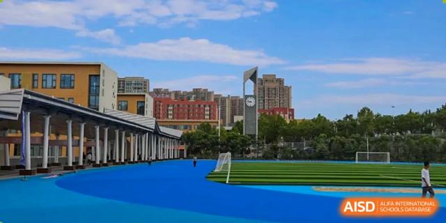 An outdoor blue running track and sports field with school buildings and a city skyline visible under a blue sky at Weiming International Qingdao.