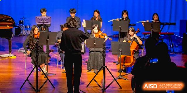 Students from Shanghai Qibao Dwight High School perform music on a stage with a conductor in front of an orchestra.