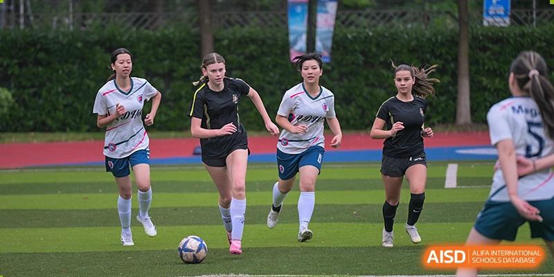 Female students from Shanghai Qibao Dwight High School run after a soccer ball on a green field.