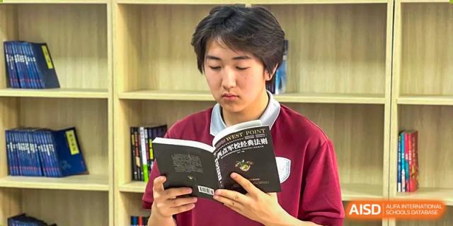 A male student in a maroon sweater reading a book intently in a quiet area or library at Shenyang Feiyue Experimental School.