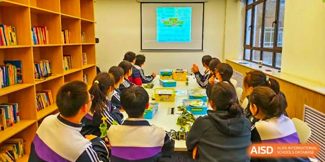 A group of students gathered around a table in a classroom, looking at a projected image during a lesson or presentation at Shenyang Feiyue Experimental School.