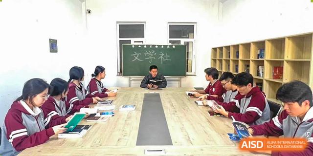 A small group of students seated around a conference table for a club meeting, possibly the "Literary Society" based on the wall text, at Shenyang Feiyue Experimental School.