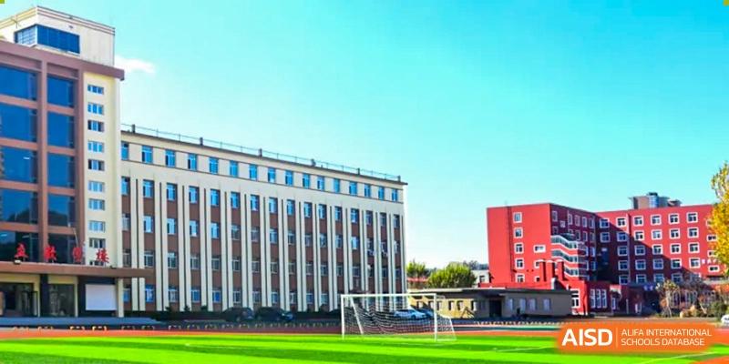 A modern school building with large windows next to a green sports field on a sunny day at Shenyang Feiyue Experimental School.