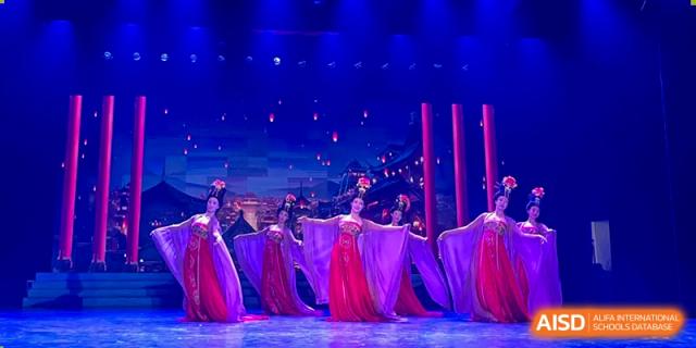 Students performing a traditional Chinese dance in elaborate costumes on a brightly lit stage at Shenyang Feiyue Experimental School.