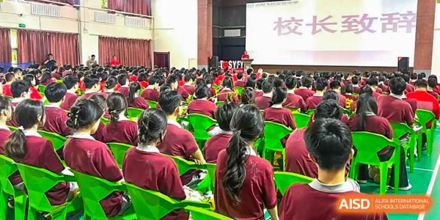 A large group of students in red shirts seated outdoors for an assembly or presentation, facing a large screen on a stage at Shenyang Feiyue Experimental School.