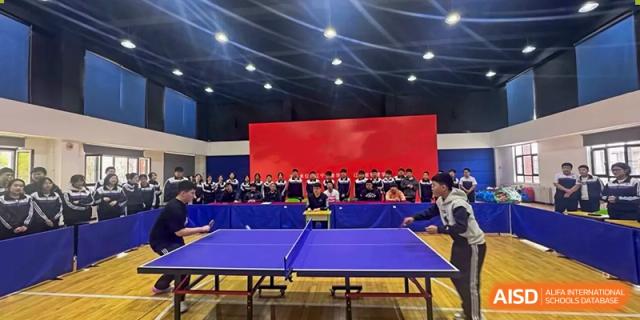 A table tennis competition or demonstration taking place in a school gymnasium with an audience seated behind a red backdrop at Shenyang Feiyue Experimental School.