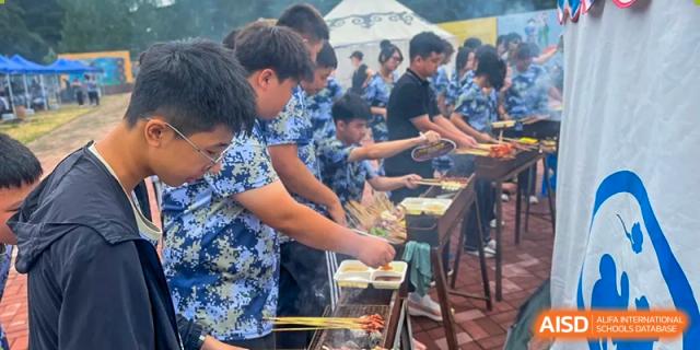 Students cooking and handing out food, likely barbecue, at an activity organized by Shenyang Feiyue Experimental School.