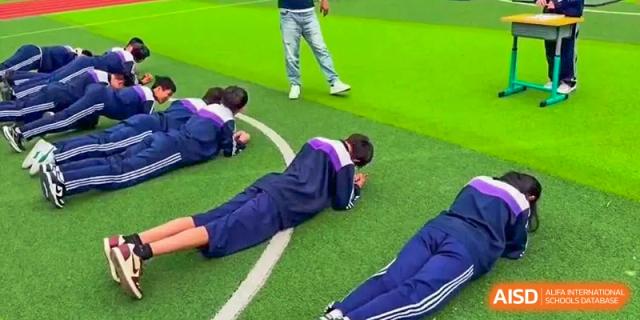 Students doing a physical education exercise, possibly a push-up or stretch, on the sports field at Shenyang Feiyue Experimental School.