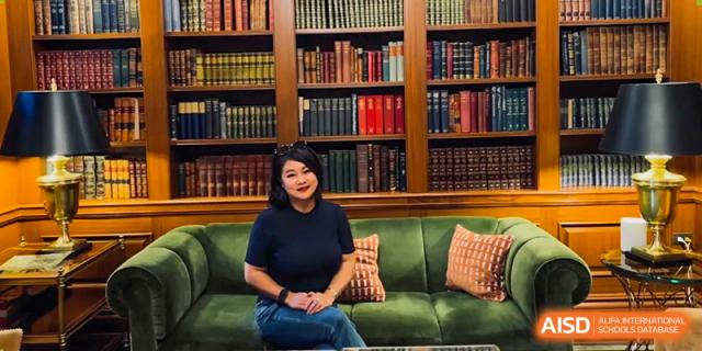 A woman is smiling while sitting on a green couch in front of a large bookshelf at Sino-Canada School.