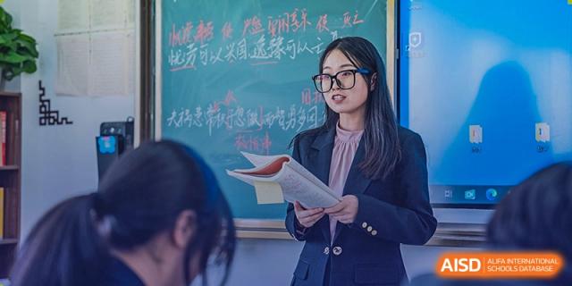 A female student from Sino-Canada School, wearing glasses, holding a book and looking at a chalkboard.