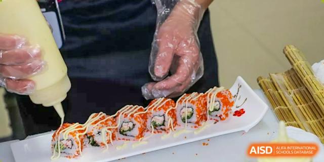 A close-up of a person wearing a plastic glove, preparing a plate of sushi rolls topped with bright orange roe and a drizzle of mayonnaise Suzhou Experimental High School International Center.