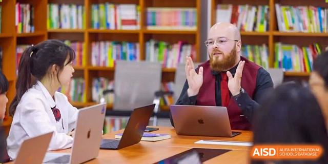 A male teacher with a beard is talking to a student while sitting in a library at The MacDuffie School Tianjin.