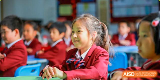 A female student in a red jacket is smiling while sitting in a classroom at The MacDuffie School Tianjin.